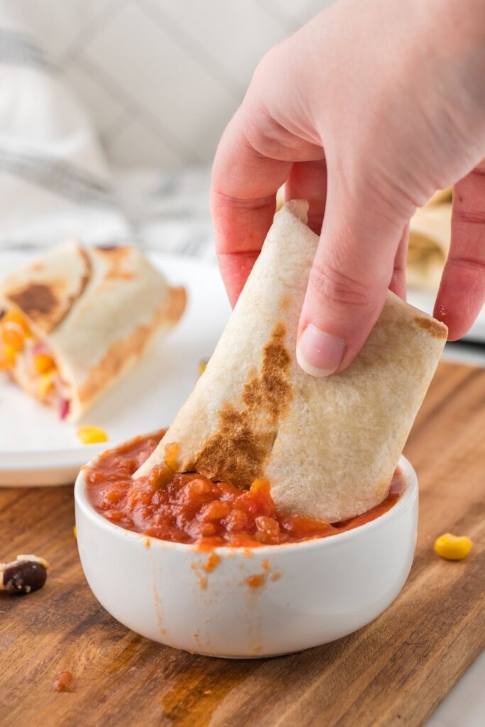 Half of a vegan burrito being dipped in salsa in a white bowl.