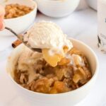 Vegan apple crisp in a white bowl with dairy free ice cream, a spoonful is being scooped up.