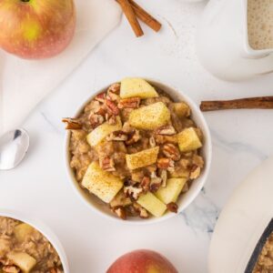 Overhead view of apple cinnamon oatmeal, with apples, cinnamon sticks, milk, and another bowl around the edges of the scene.