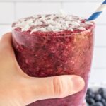 Hand holding berry smoothie with coconut water topped with coconut flakes with a blue striped straw.