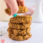 Hand with teal painted nails picking up a peanut butter chickpea cookie from a stack of cookies.