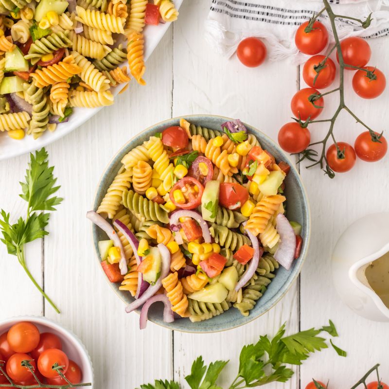 Overhead view of pasta salad in a blue bowl with the white serving tray at the top left and other ingredients surround the main bowl.