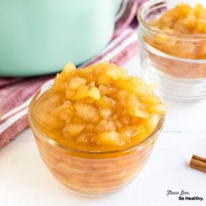 Simple homemade applesauce in a small glass bowl with a teal pot and another bowl of applesauce behind it.