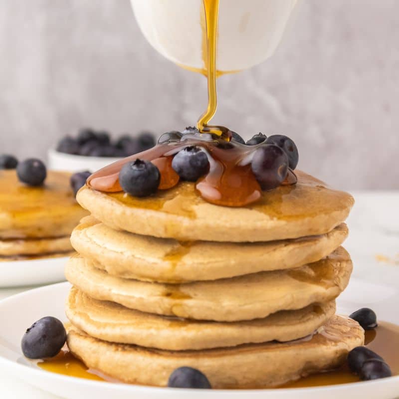 Maple syrup being poured from a white pourer onto fluffy vegan oatmeal pancakes with blueberries on top. These easy vegan oatmeal pancakes have no egg, no banana, and are easy to make.