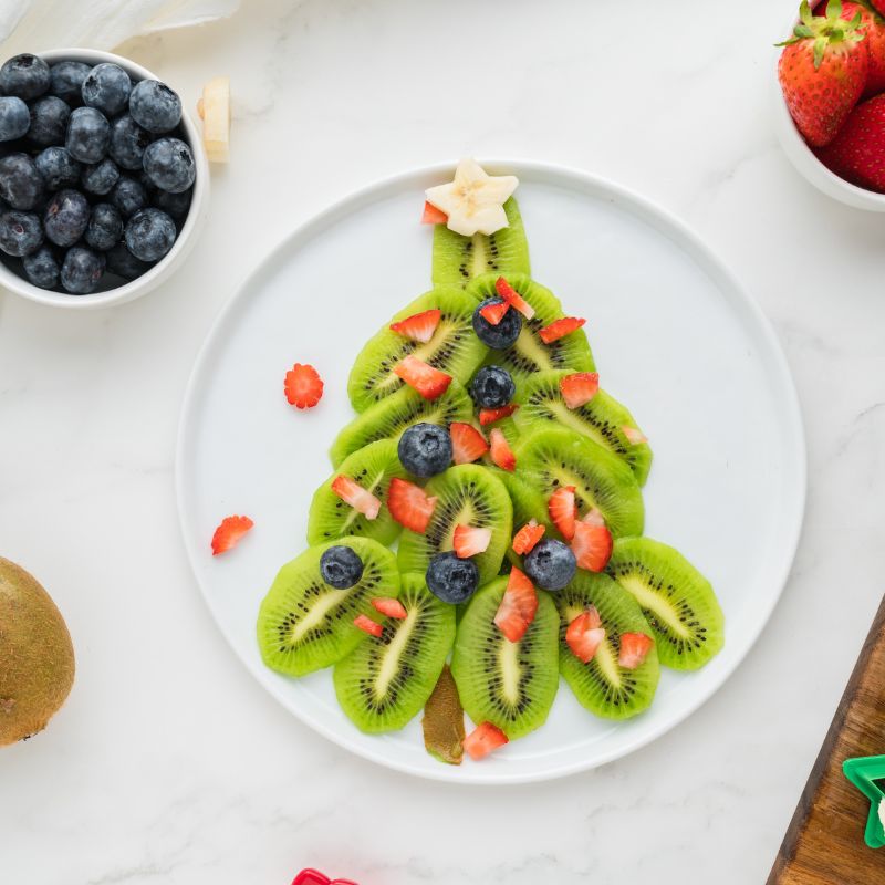 Kiwi Christmas tree with blueberry and strawberry ornaments and a banana star tree topper on a white plate with more ingredients surrounding the tree.