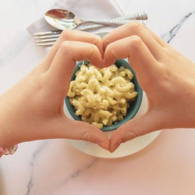 Easy vegan macaroni and cheese in a blue bowl, overhead photo, with kids hands making a heart above the bowl of delicious pasta.