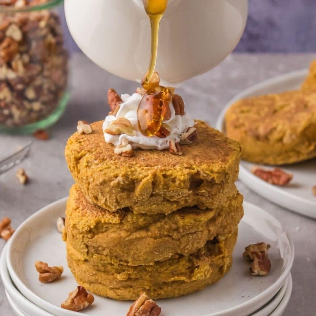 Vegan Pumpkin Pancakes with non-dairy whipped cream and pecans on top, with maple syrup pouring over it.