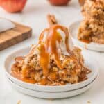 Single bar of vegan apple pie bar recipe with vegan vanilla ice cream and caramel sauce drizzled on top, all on a white plate, with more bars and apples in the background.