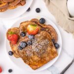3 slices of Vegan French Toast on a white plate with berries and powdered sugar on top.