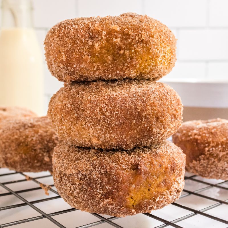 3 vegan pumpkin donuts with cinnamon sugar stacked on a cooling rack, with more donuts and non-dairy milk behind the stacked donuts.