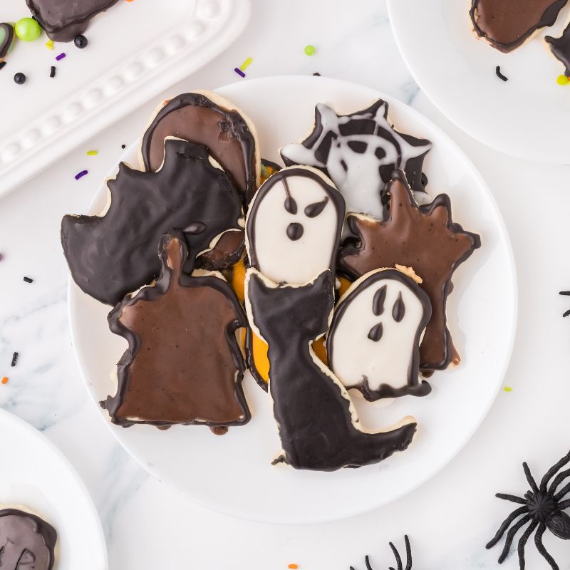 Vegan halloween sugar cookies in the shapes of cats, ghosts, haunted house, bats, old spooky tree, and spider web all iced and on a white plate, with fake spiders near it.