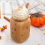 Iced Pumpkin Spice Latte in a medium sized mason jar with vegan whipped cream, cinnamon stick, and some extra pumpkin pie spice sprinkled on top, with a glass straw.