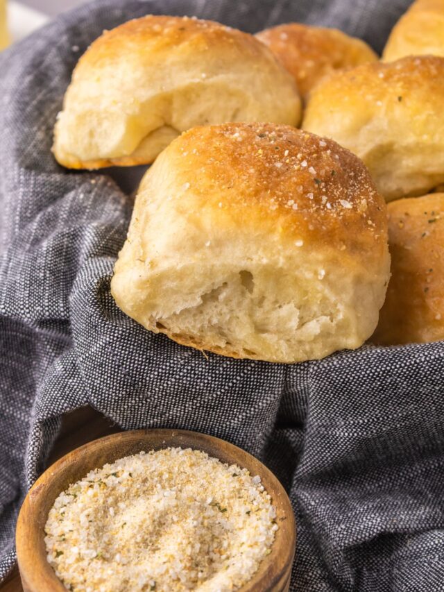 Vegan dinner rolls in a bread basket with a blue-gray linen underneath and garlic salt next to it.