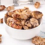 Candied Pecans in a white bowl with more pecans surrounding it.
