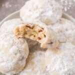 Vegan Snowball cookies piled on a white plate with the top cookie with a bite missing.