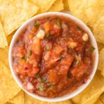 Homemade salsa in a white bowl in the middle of a bunch of tortilla chips.