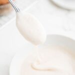 Vanilla icing drizzling down from a spoon into the white bowl it was made in.