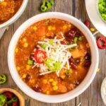 Sweet potato quinoa chili served in a white bowl with sliced tiny sweet peppers, green onions, and vegan shredded cheese.