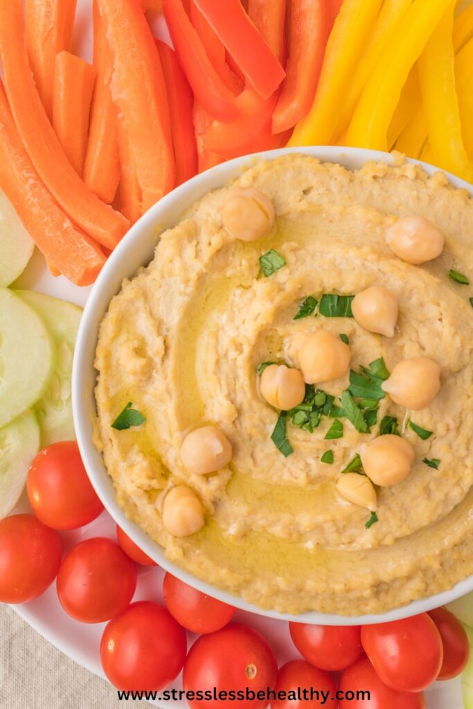 Homemade hummus in a white bowl with parsley, olive oil, and whole chickpeas added to the top surrounded by colorful raw veggies.