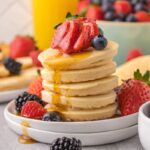 Mini pancakes stacked on a white plate with berries and syrup on top and on the plate.