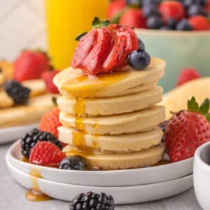 Mini pancakes stacked on a white plate with berries and syrup on top and on the plate.