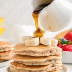Stacked vegan banana pancakes with extra banana slices on top and syrup being poured on top, with bright red berries in the background.