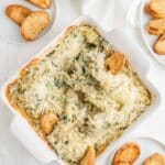 Vegan spinach artichoke dip in a white square baking dish with baguette and crostini in it and around it.