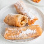 3 apple pie spring rolls on a plate, covered in sifted powdered sugar, middle one has a bite taken out of it to show the inside.