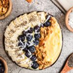 Mango smoothie bowl topped with fruit, coconut shreds, and granola, with extra ingredients surrounding the bowl.