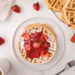 Vegan Strawberry waffles on a white plate covered in strawberry syrup and powdered sugar.