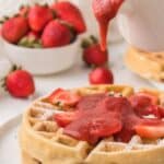 Strawberry waffles with strawberry syrup being poured on it, with more strawberries in a bowl in the background.