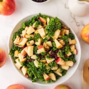 Kale Apple Salad in a white bowl surrounded by dressing and apples.