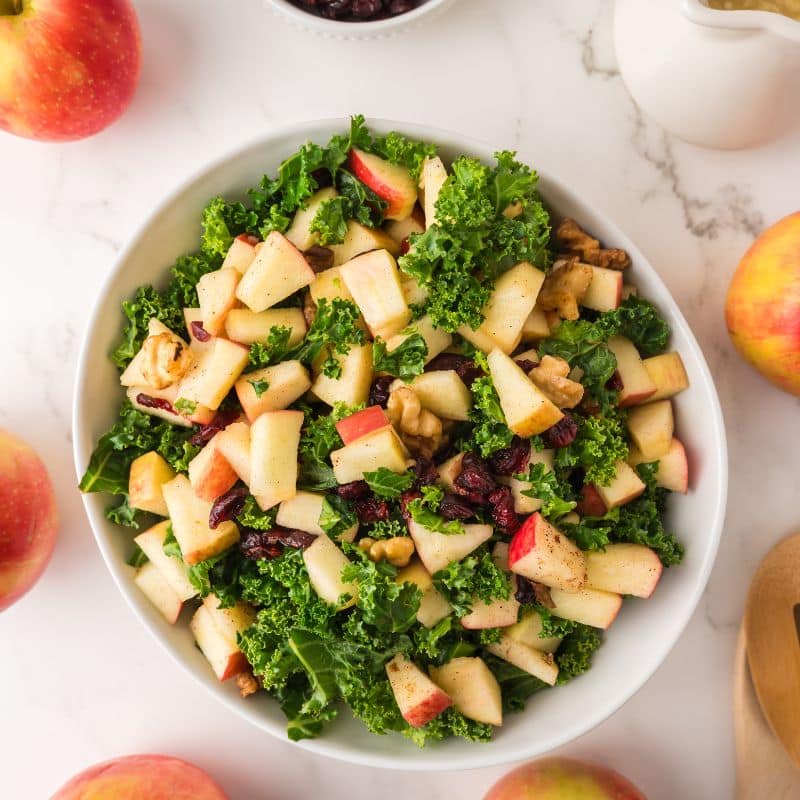 Kale Apple Salad in a white bowl surrounded by dressing and apples.