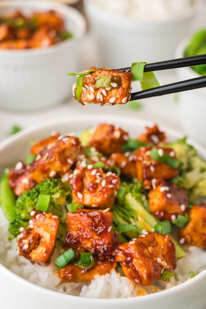 Gochujang tofu in bowl with broccoli, sweet peas, and white rice, with one piece being held up by black chopsticks.